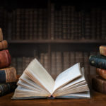Opened,book,and,stacks,of,old,books,on,wooden,desk