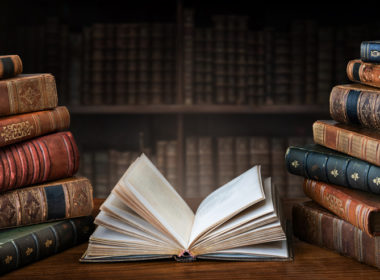 Opened,book,and,stacks,of,old,books,on,wooden,desk