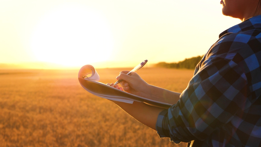 Farmer,writing,notes,in,notebook,against,golden,wheat,field ,close up